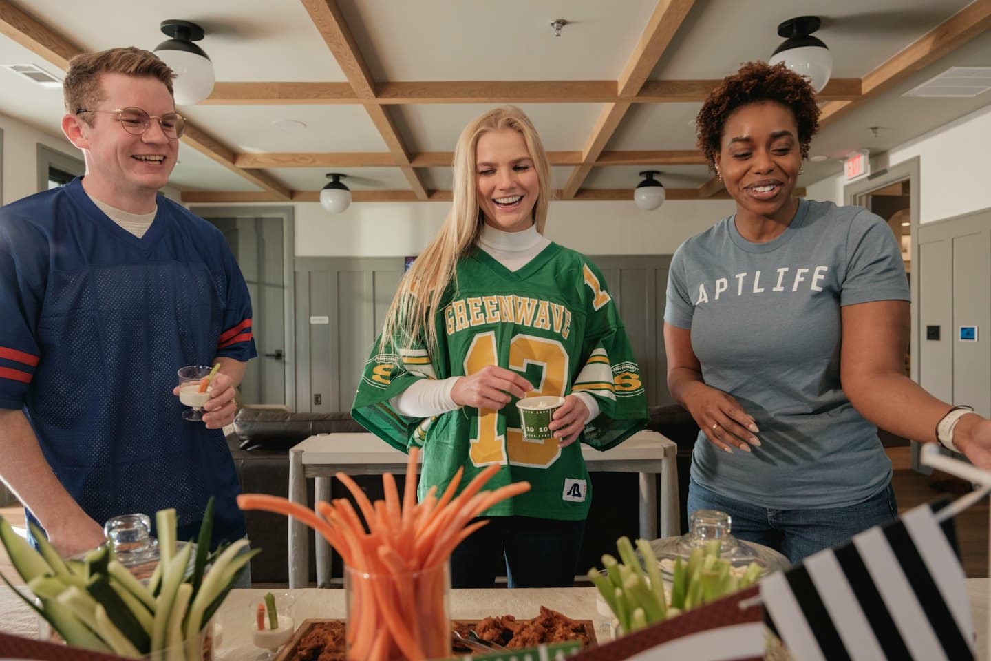 Three friends stand around a table sharing food and drinks.