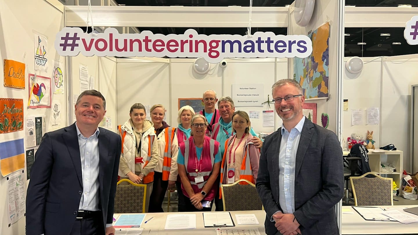 Two men in suits stand with volunteers at an exhibition booth.
