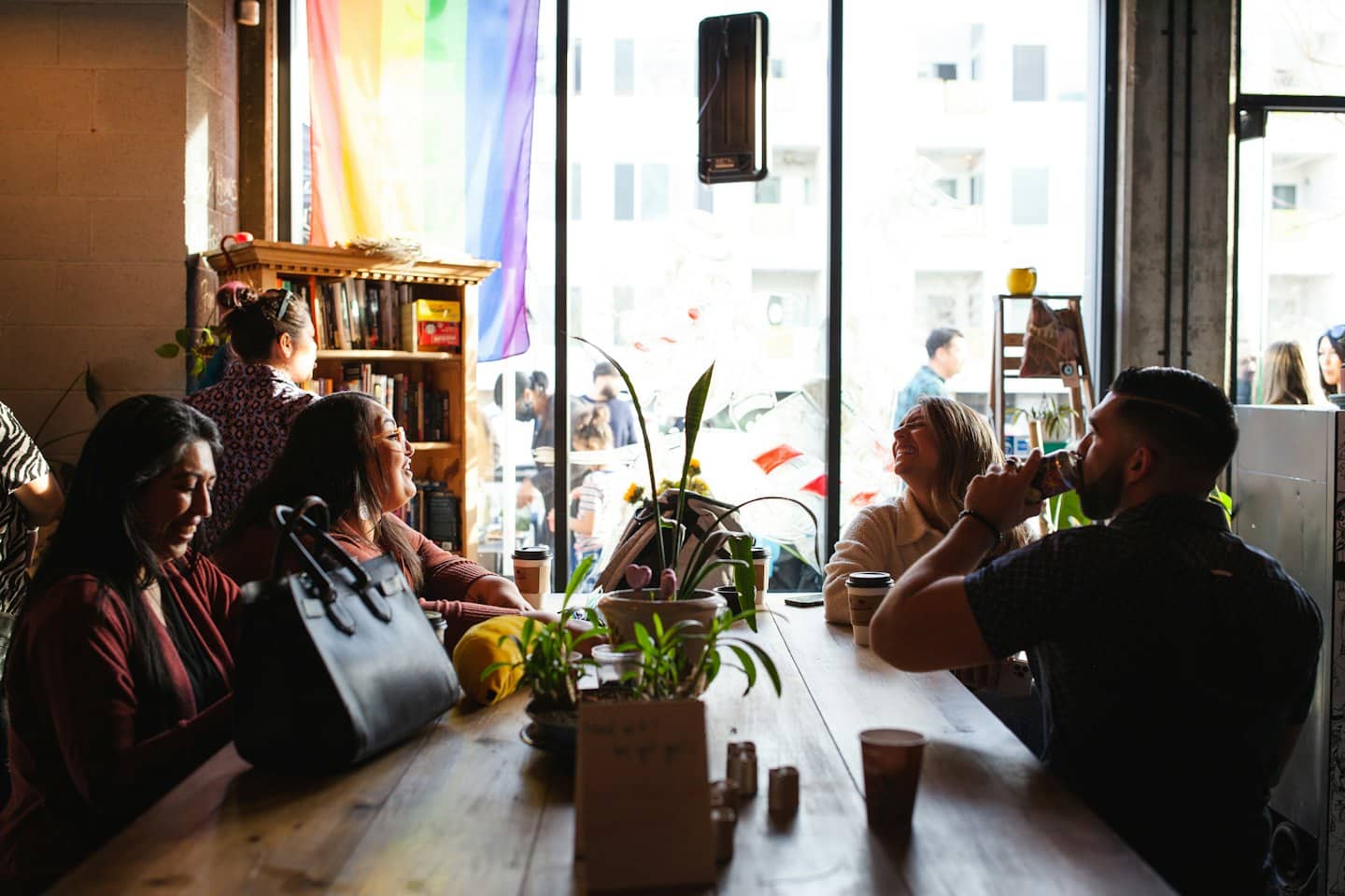 Diverse group laughs at a cafe table with plants and coffee.
